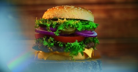 A big tasty burger with meat patty, onions, vegetables, melted cheese, lettuce and mayonnaise sauce. Isolated hamburger rotates on a dark wooden background.