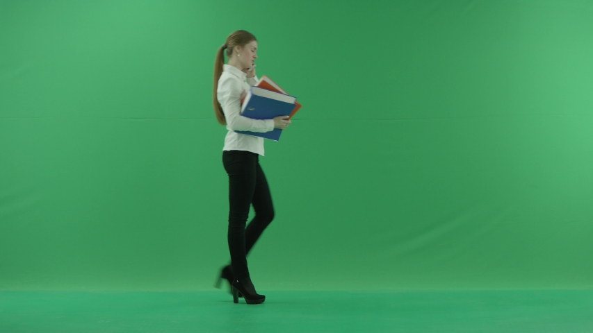 business woman is talking on her mobile cell phone and walking with folders. She wears formal dress: white shirt and blak trousers. Businesswoman is over green screen