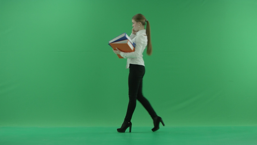 business woman is talking on her mobile cell phone and walking with folders. She wears formal dress: white shirt and blak trousers. Businesswoman is over green screen
