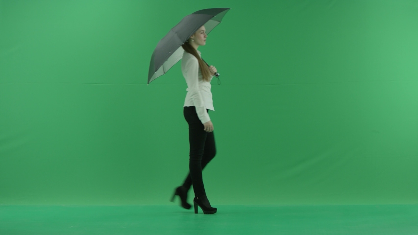 a thoughtful woman is walking right side with umbrella over green screen . She wears formal dress: white shirt and blak trousers.