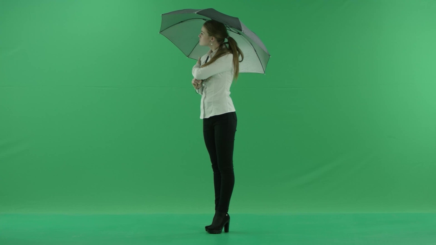 a thoughtful woman is waiting standing left side with umbrella over green screen . She wears formal dress: white shirt and blak trousers.