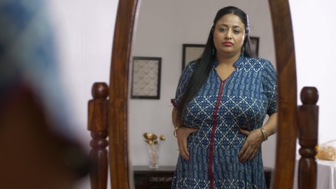Indian overweight woman pinching her belly fat with frowning gesture - weight control concept. Reflection of a beautiful mature lady checking her belly fat looking into the mirror - Unfit woman