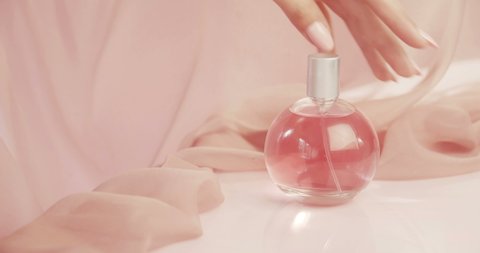 Glass perfume bottle with pink liquid stands on table. Pink silk fabric flutters around. Beautiful elegant female hand with pink nails and long fingers touch bottle of perfume. Concept for perfume.