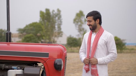 Young attractive farmer happily standing beside his brand new tractor. Handsome Indian villager purchased a new tractor for his farm, giving a wide smile while folding his hands - agricultural fiel...