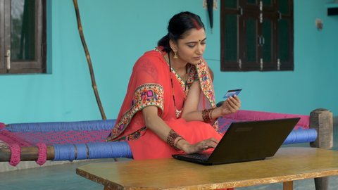 Young beautiful village woman doing card payment - online shopping concept. Rural life. Attractive Indian villager holding debit/credit card in her hand and entering card details with another hand ...