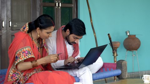 Indian village couple doing card payment using laptop - online shopping concept. Young beautiful wife holding a debit/credit card in her hand and telling her husband card details to do online payment