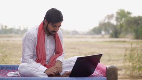 Educated Indian farmer learning new things on a laptop - technology concept. Young villager using laptop and internet for the first time while sitting on a charpai - Indian agricultural field in th...