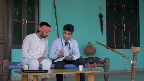 Young Indian farmer getting information about health insurance policy from an agent. Happy village man listening to well-dressed man. Sitting on a charpoy with desi hukka. Village Home