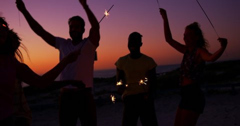 Close up of a multi-ethnic group of young adult friends on a beach at sunset, dancing around and having fun waving glowing sparklers. Summer Sunset Party on the beach with friends having fun 4k