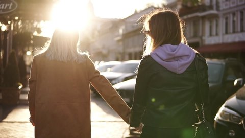 Two happy young girls walk by the city together, hug each other. Females embracing, laughing and excited. Woman friendship, walk in the city outdoors. City view, sunlight on the background