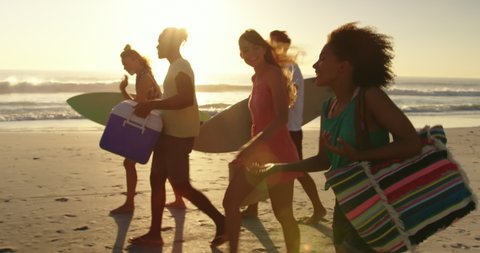 Close up side view of a multi-ethnic group of happy young adult friends walking on a beach at sunset, carrying surfboards, a cool box and beach bags. Backlit. Summer fun with friends, they are on a