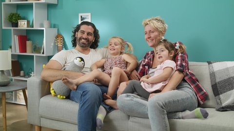 Happy family watching tv. Couple with daughters sits on a gray sofa and laughs.
