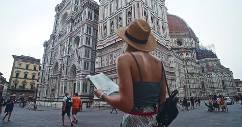 Florence, Italy - 18 June, 2019: Pretty girl tourist observing large majestic Duomo Cathedral. Young woman in focus holding city map against stunning Santa Maria del Fiore. Piazza del Duomo.