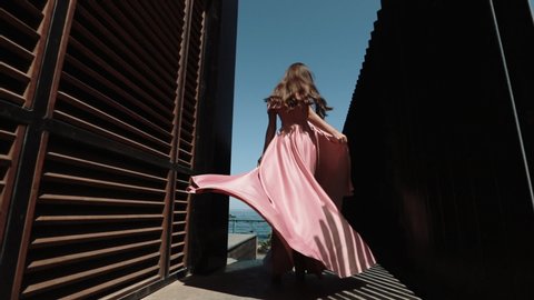 Following Shot Of Attractive Woman Relaxing Near Seaside Building On Popular Tropical Resort During Paradise Island Vacation. Beautiful Fashion Model In Pink Luxury Formal Dress Is Posing On Balcony.