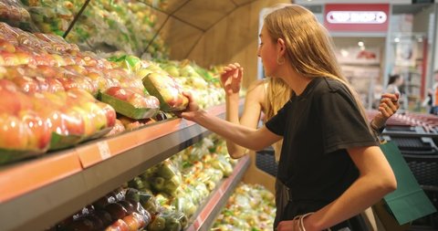 Portrait of two pretty girls when they choose apples in the supermarket. Two young women choosing a dairy products, vegetable, fruits at the supermarket. 4K video