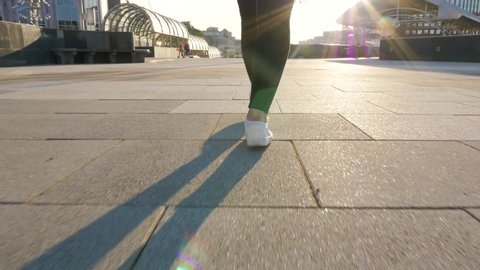 female legs in black trousers and white shoes walk along a clean, empty path. towards the sun, shooting from a low point, the camera follows the movement. city walk in slow motion close up