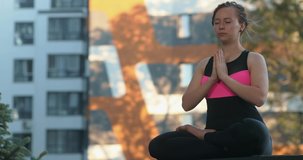 Young woman in sportwear is sitting in the lotus pose and meditating. He is doing yoga exercises outdoors on balcony. 4K video