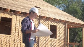 concept building constructing architect slow motion video. man builder in a helmet stands at construction holding a scheme house plan. site near a lifestyle wooden frame house under construction