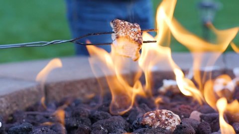 
A backyard tradition as roasting marshmallow on a campfire. Slow motion close up shot as someone is holding a skewer stick with white roasted marshmallow over open fire place in green backyard.