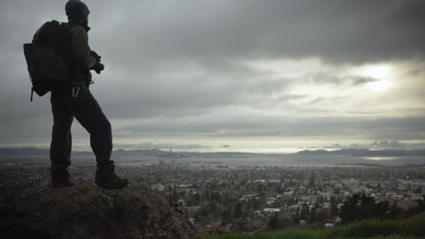 A photographer proudly stands on a cliff with an incredible view of the city and sunset on a beautiful cloudy evening.  Shot in 4K UHD resolution.