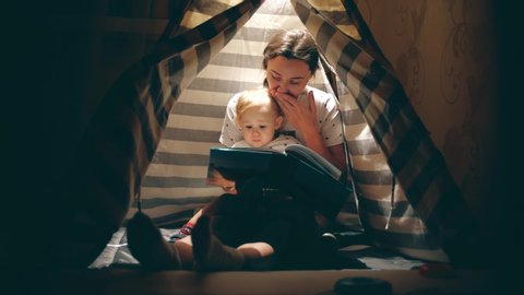 Mother and her little baby read a book together in a cozy lit teepee in the evening