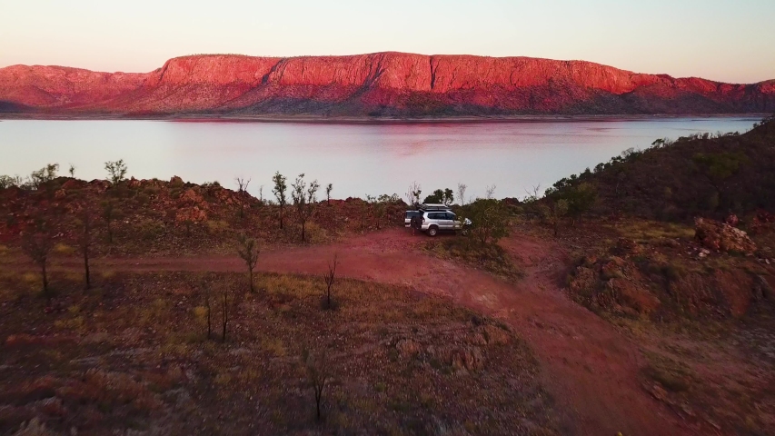 Spectacular fast aerial fly-over of car on mountain top beside huge Lake at sunset with red mountains in background. Location Lake Argyle, Australia.