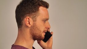 Red-haired man is standing sideways and chatting on the phone, white background. 4k video