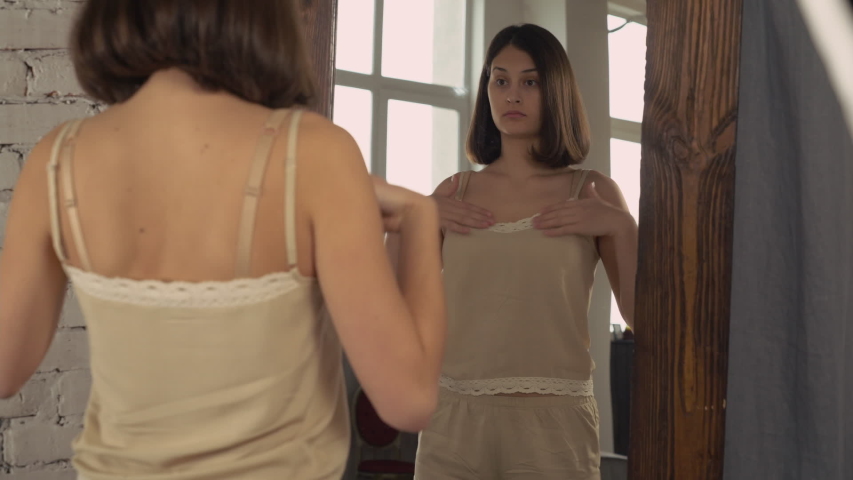 Woman looking in the big mirror. Young lady stands in bedroom wearing in pajamas shorts and top nude color. Happy girls in the morning.