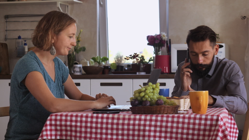 Small business partners have meeting at home. woman working on laptop. man has phone negotiation using smartphone. Young people sitting at the table in kitchen | Shutterstock HD Video #1034410541