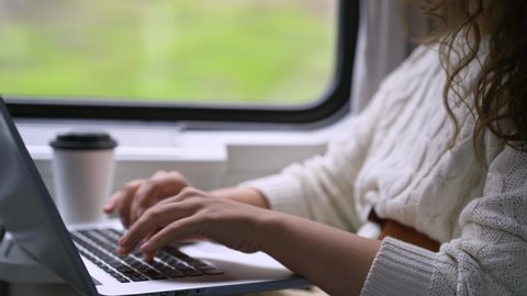 Young girl travelling on train in daytime. Woman sitting near window, working on computer. Detail view female hands typing on laptop keyboard and taking white paper cup with coffee