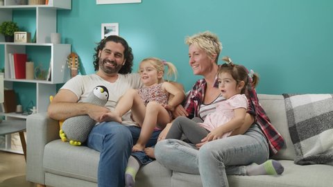Couple with daughters sits on a gray sofa watching tv and laughs.