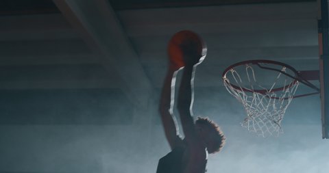 Cinematic slow motion shot of an young african professional male player is making a  slam dunk during a basketball work out in a gym.