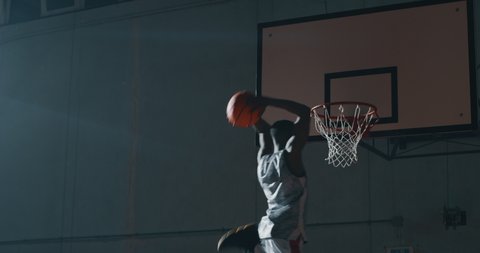 Cinematic slow motion shot of an young african professional male player is making a  slam dunk during a basketball work out in a gym.