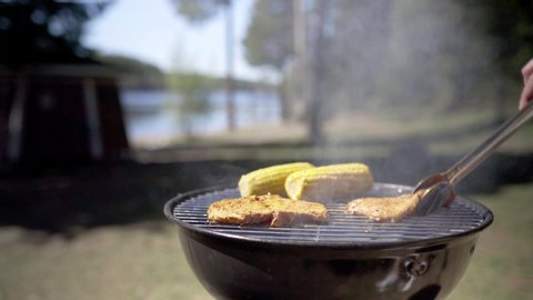 Delicious food in smoking outdoor grill. Woman cooking for BBQ party. Kitchen in nature. Barbeque cookout. Charcoal kettle by the lake or in park or forest. Sunny summer day.