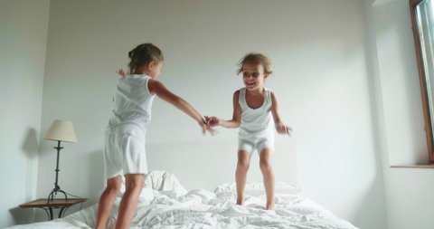 Authentic slow motion shot of two little girls sisters in white pajamas are having fun to jump on the bed of their parents bedroom.