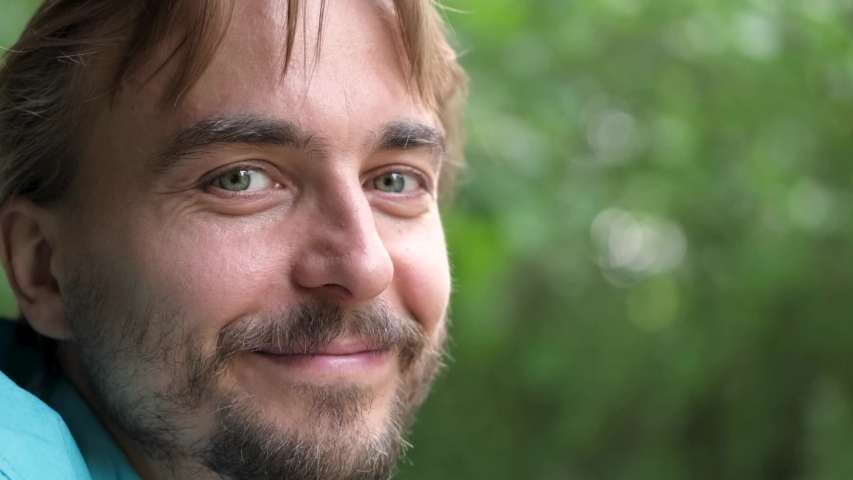 Handsome bearded caucasian man with green eyes turns to the camera and smiles. Slow motion. Blurred nature background. | Shutterstock HD Video #1034430716