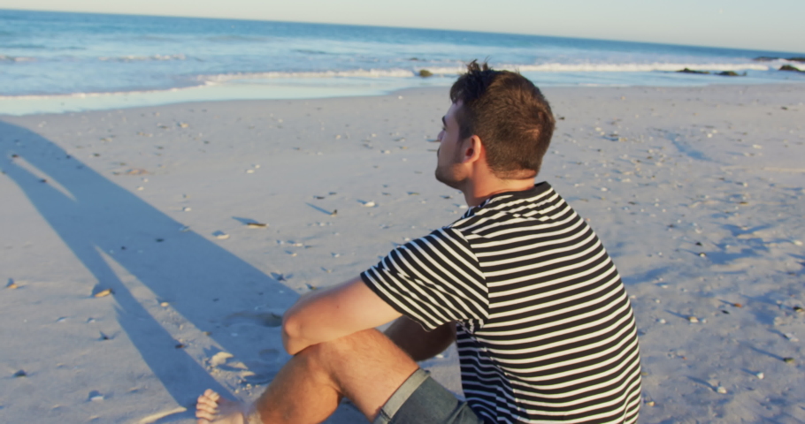 Close up of a young Caucasian man siting on a beach looking out to sea, turning and smiling to the camera 4k