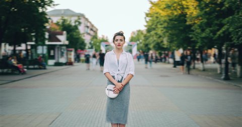 Zoom in time lapse of serious young woman in casual clothing standing alone in busy street looking at camera while people men and women walking around.