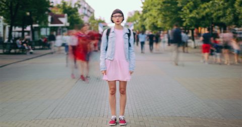 Time lapse of attractive teenage girl standing in city center in busy street looking at camera wearing trendy clothes while crowds of people are walking by.