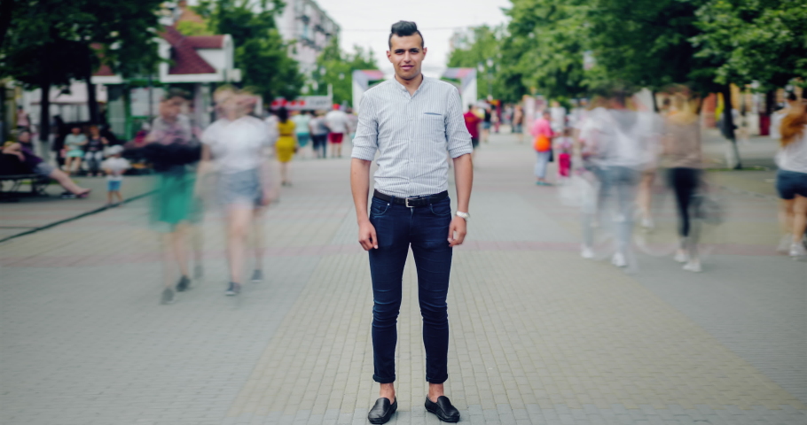 Time lapse portrait of handsome Arabian man in casual clothing outdoors in city street standing alone with serious face looking at camera while people moving around.