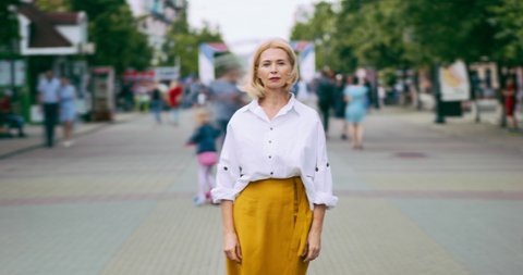 Zoom out time lapse of pretty mature woman looking at camera standing in street metropolis with serious face wearing elegant clothing while people are rushing around.