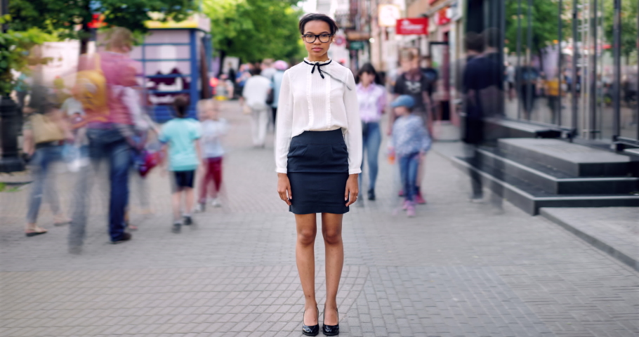 Time lapse portrait of attractive African American girl in elegant clothing standing in city street with serious face looking at camera while people are walking around. | Shutterstock HD Video #1034434730