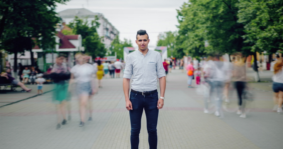 Zoom in time lapse portrait of Arabian student standing in street among flow of people looking at camera with serious face. Youth, modern life and summer concept.