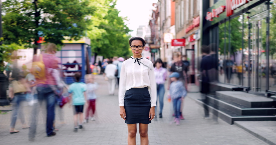 Zoom in time lapse of pretty African American lady standing in busy street on summer day looking at camera with serious face. Modern lifestyle, people and youth concept.
