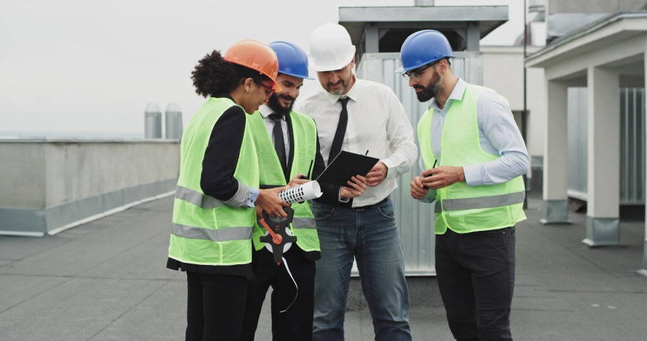 Charismatic young African lady multiethnic group of workers on the top of construction site they chatting with each other analyzing the plan of building wearing the hard hat safety helmets