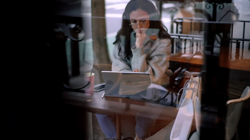 Window view of attractive millennial woman in optical spectacles making online messaging with virtual friends during distance job on portable pc, Caucasian female using touch pad for chatting