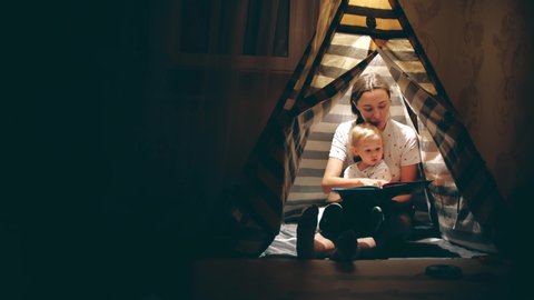 Mum and her little daughter read a book together in a teepee in the evening