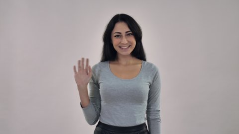 happy adult mixed race caucasian woman with black straight hair greeting showing symbol hi. Portrait female wearing casual grey shirt posing in studio on background