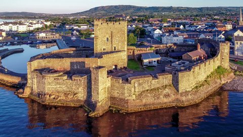 Medieval Norman Castle and harbor in Carrickfergus near Belfast in sunrise light. Aerial 4K revealing video with water reflection, marina, breakwater, town and far view of Belfast in the background