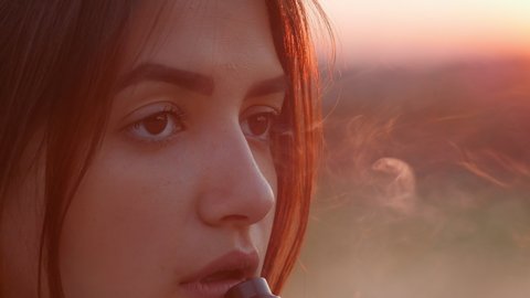Cute closeup face portrait of relaxed young girl vaping e-cigarette outdoors with cool red sunset light